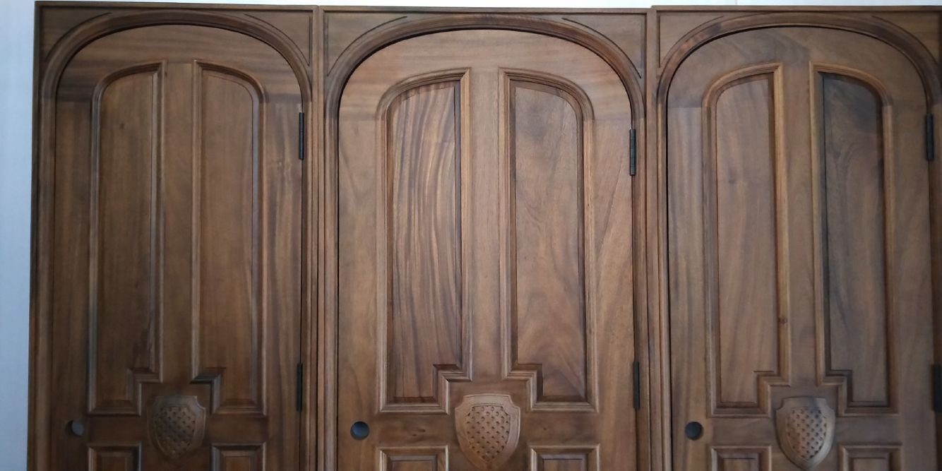 A detailed architectural view of three finished, custom arched solid wood doors from the file crown-custom-doors-arched-black-walnut-doors-shield-emblem.jpg, handcrafted from premium solid black walnut with a deep satin finish, featuring rich organic wood grain, complex stepped panel molding, and a detailed hand-carved shield emblem at the base of the main panels.