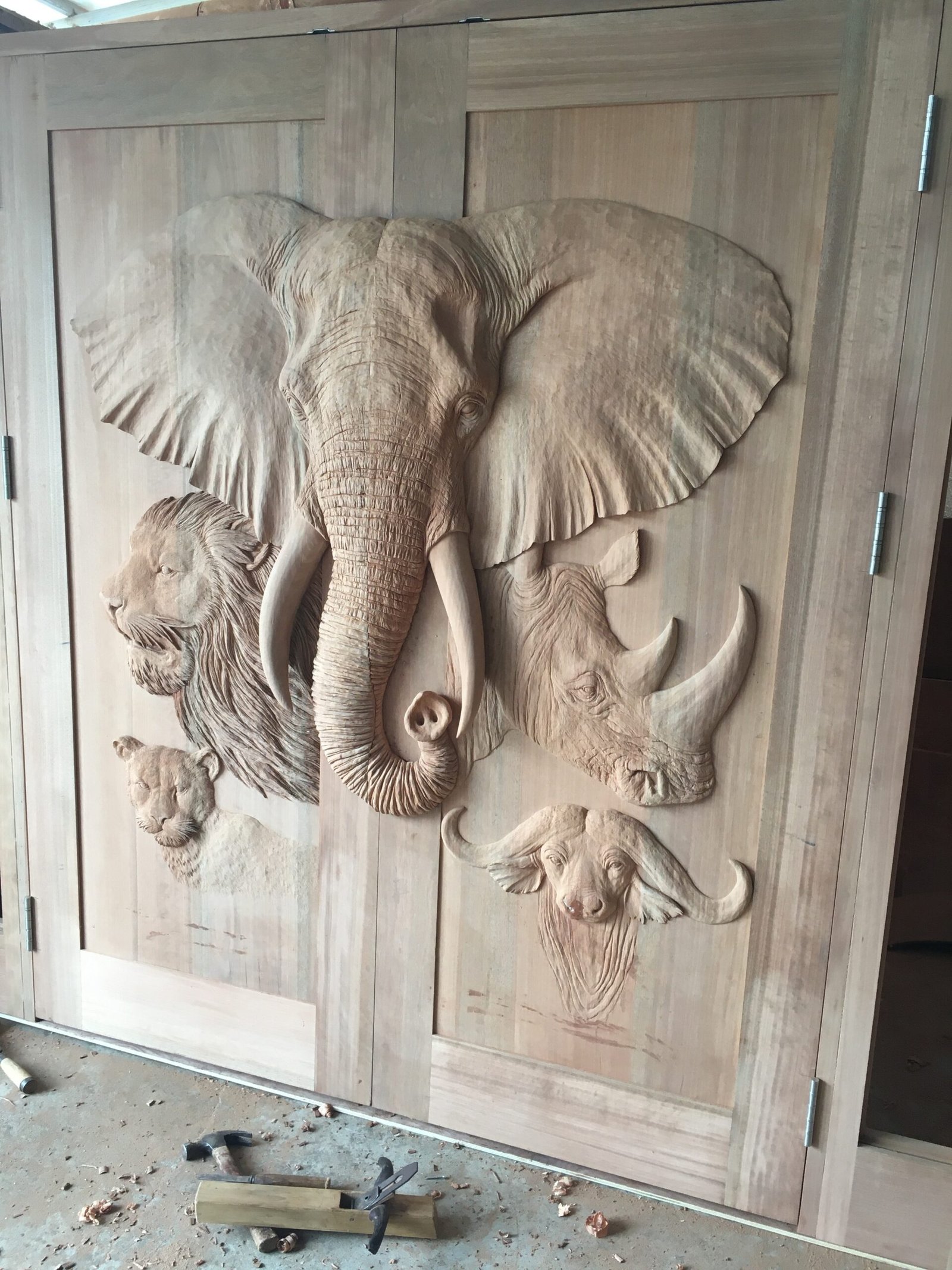 An angled, three-quarter workshop view of a raw solid wood double door showcasing the deep 3D dimension of the hand-carved Wild Africa safari animals, including the prominent elephant head, lion, rhinoceros, leopard, and cape buffalo with woodcarving tools on the floor.