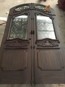 Full view of a custom arched double wood entry door lying flat during assembly in a workshop, featuring raised bottom panels, hand-forged wrought iron grilles, and black cremone bolt locks running down the center.