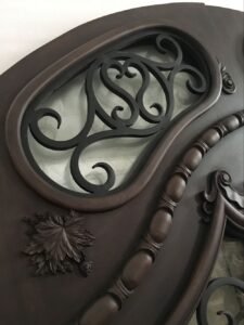 Close-up of the upper arched transom on a custom solid wood entry door, featuring a curved glass insert with black wrought iron scrollwork, a hand-carved rope molding, and a detailed botanical leaf carving.