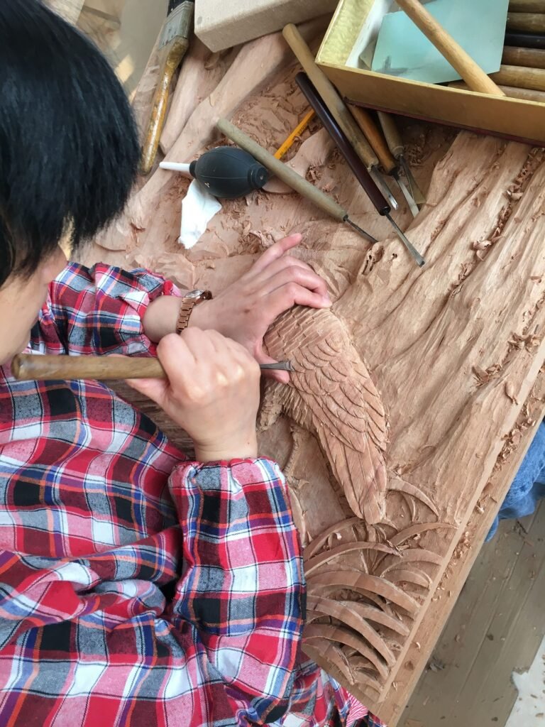 Overhead view of a master woodcarver using traditional hand gouges and chisels to detail bird feather textures on a solid wood door panel.