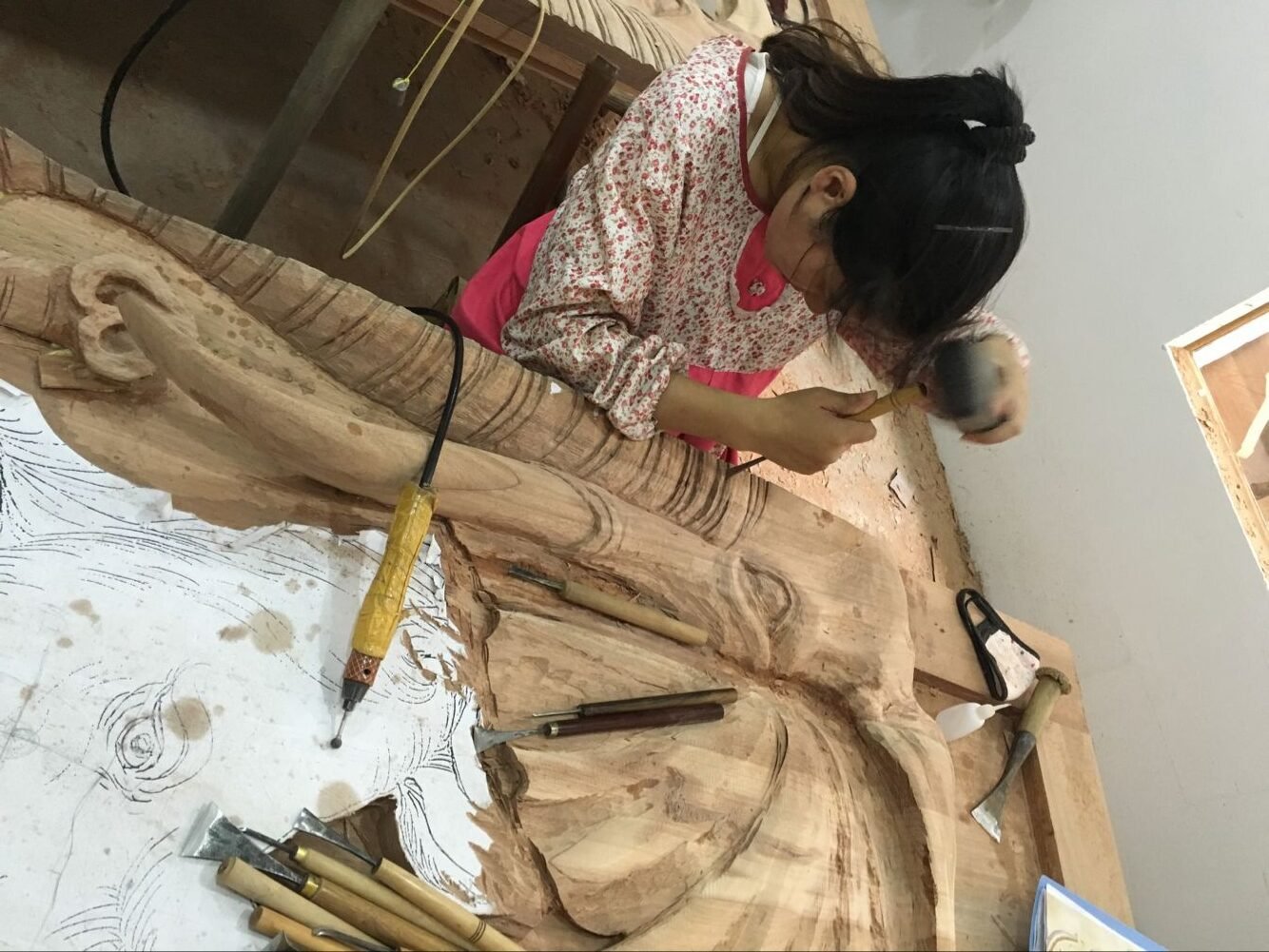 A woodcarver using a mallet and traditional wood chisel to carve high-relief details into a solid wood elephant door panel, surrounded by assorted hand chisels, fine detailing tools, and paper sketches in a custom woodworking workshop.