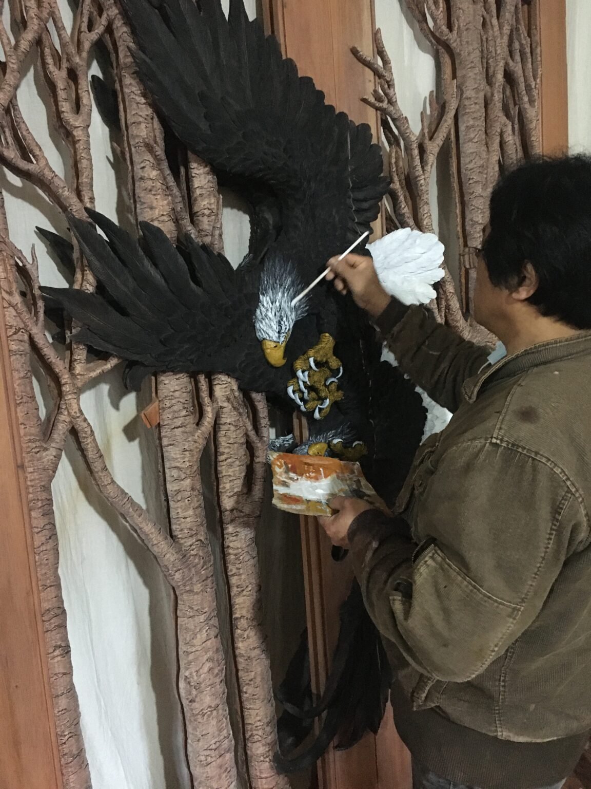 Artisan in a workshop using a fine paint brush to apply detailed white feathers and color highlights to a hand-carved solid wood eagle door.