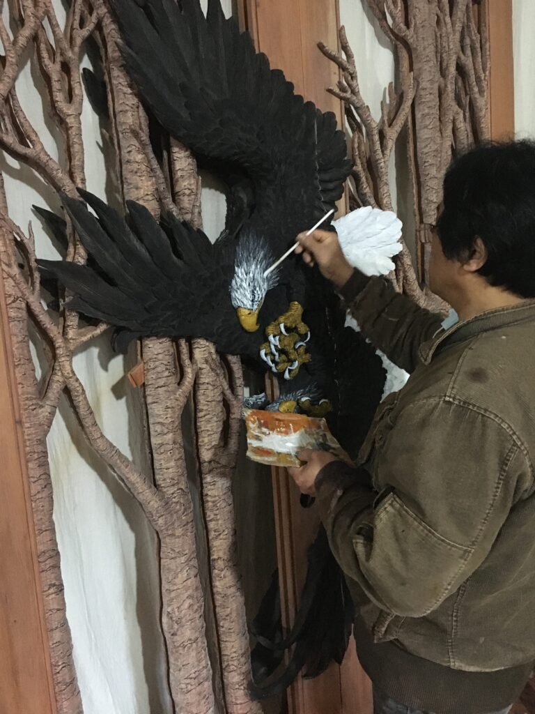 Artisan in a workshop using a fine paint brush to apply detailed white feathers and color highlights to a hand-carved solid wood eagle door.