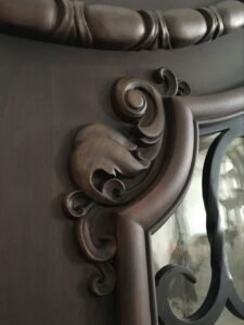Close-up of a custom solid wood door panel showing a deep, three-dimensional hand-carved acanthus leaf and baroque scrollwork molding next to a wrought iron glass panel insert.