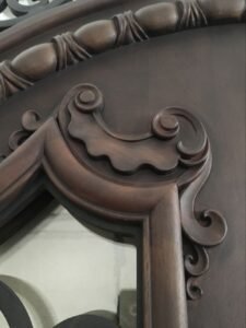 Close-up detail of a custom solid wood door showing a deep, three-dimensional hand-carved baroque scroll and scalloped wooden crest next to a decorative rope-style carved molding frame.