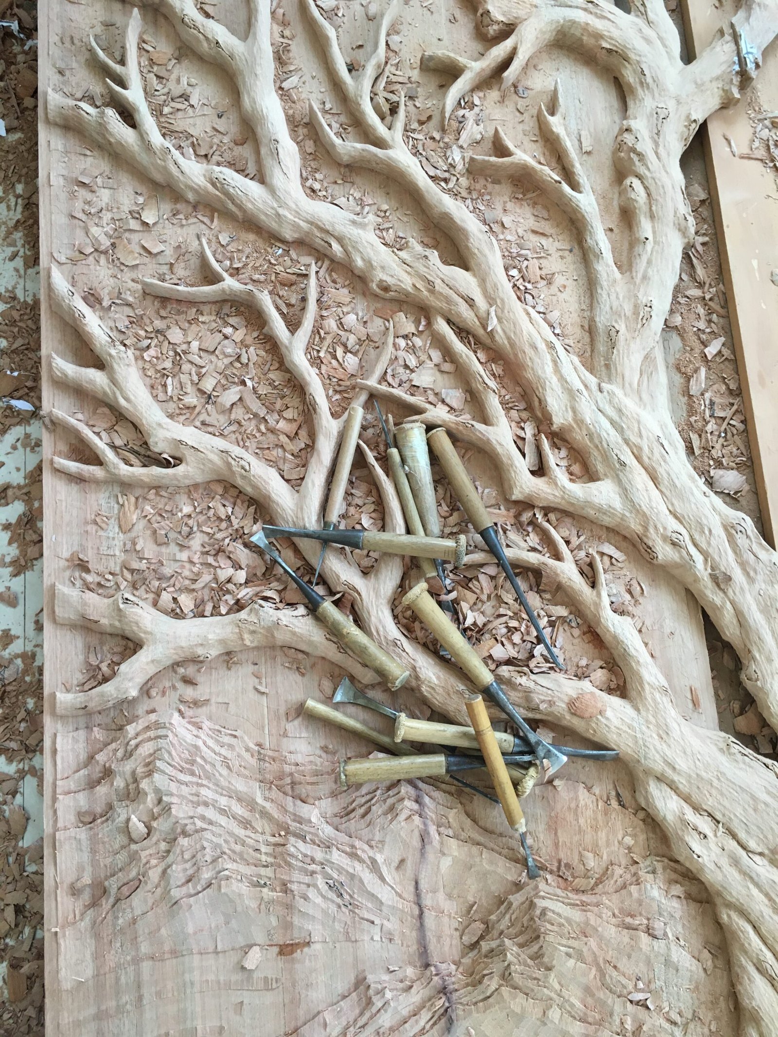 Close-up view of traditional woodworking chisels lying on hand-carved tree branches and high-relief mountain textures during the custom door fabrication process at Crown Custom Doors.