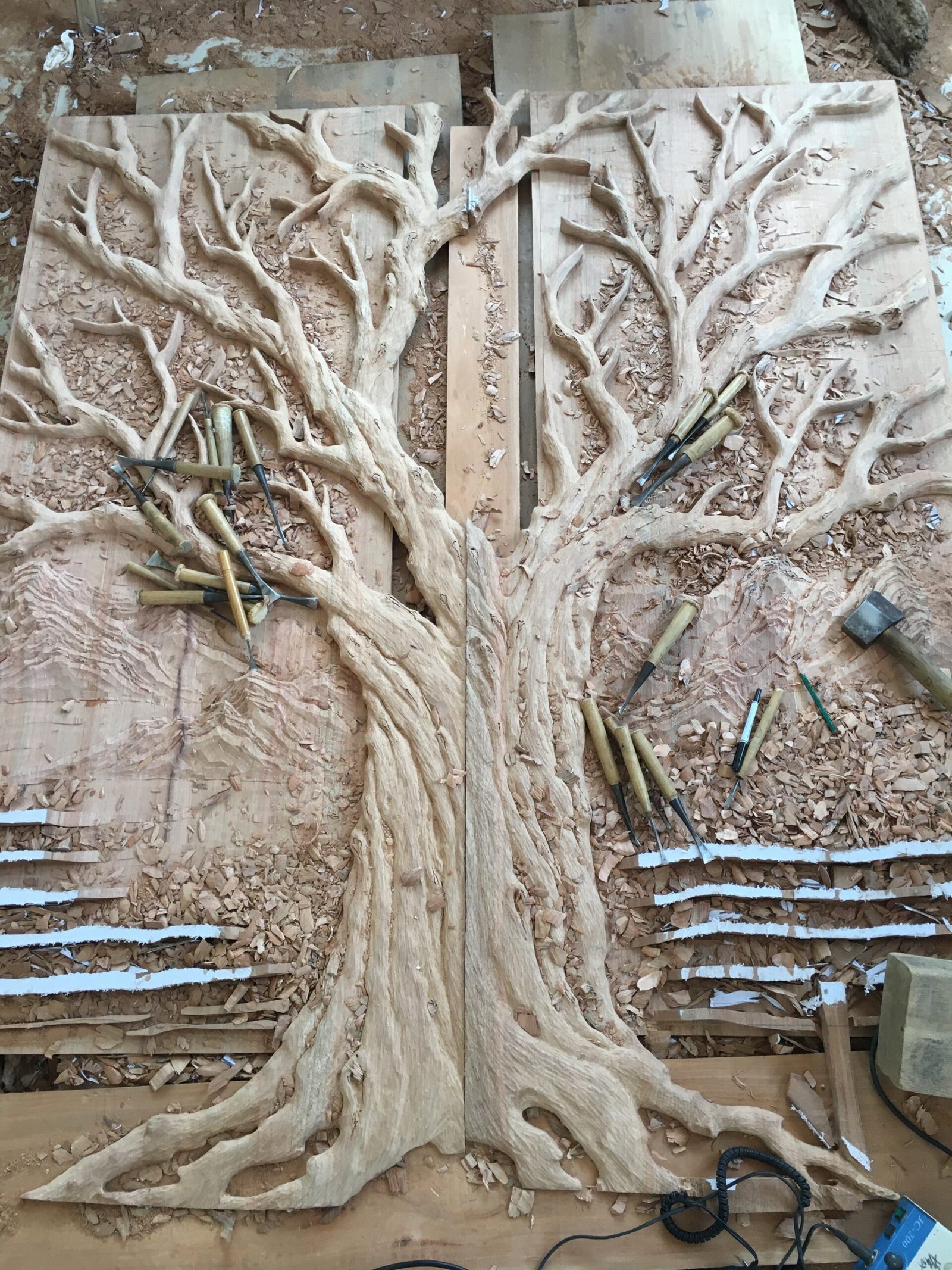 Detailed artisan hand-carving process of a solid wood Tree of Life double entry door at the Crown Custom Doors workshop, showcasing traditional woodworking chisels, wood shavings, and relief-carved mountain scenery.