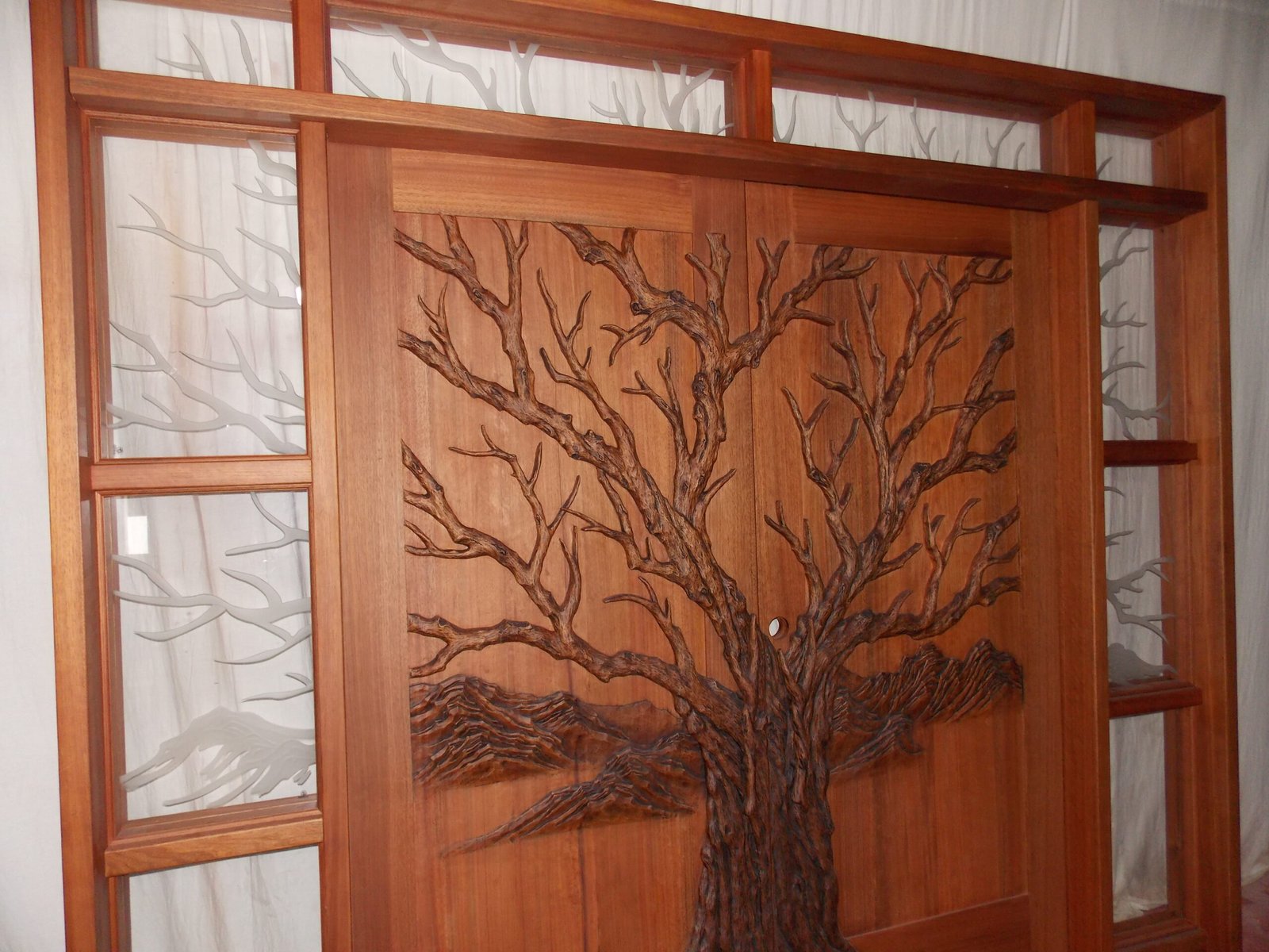 Detailed angle view of a stained solid wood double entry door by Crown Custom Doors featuring a high-relief hand-carved Tree of Life design, accompanied by frosted etched branch art glass sidelights and transom panels.