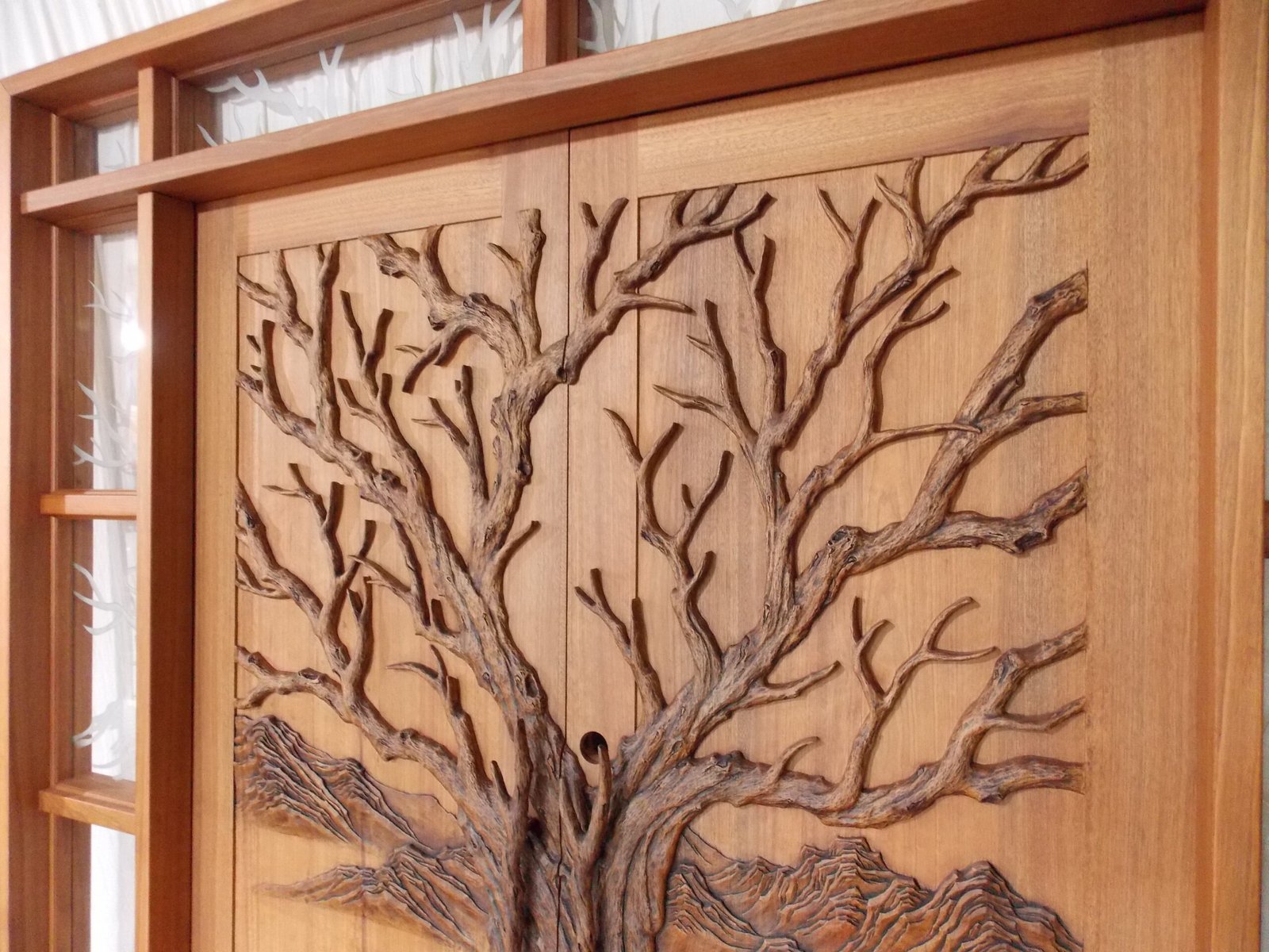Angle view of a stained solid wood double entry door by Crown Custom Doors featuring a high-relief hand-carved Tree of Life motif assembled inside a custom wood jamb frame with sidelight and transom glass inserts.