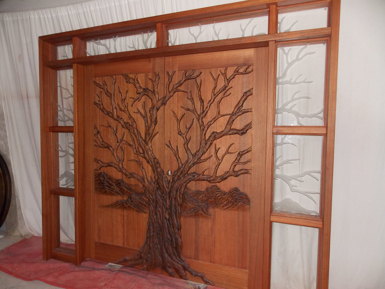 Three-quarter studio view of a stained solid wood double entry door system by Crown Custom Doors featuring a high-relief hand-carved Tree of Life design, surrounded by a custom jamb frame with etched branch sidelights and transom glass.