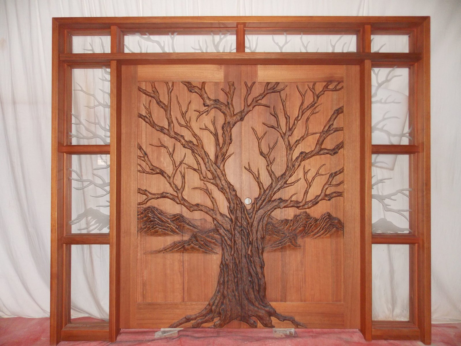 Full front studio view of a stained solid wood double entry door system by Crown Custom Doors featuring a high-relief hand-carved Tree of Life design, matching jamb frame, multi-lite sidelights, and a top transom with etched branch art.