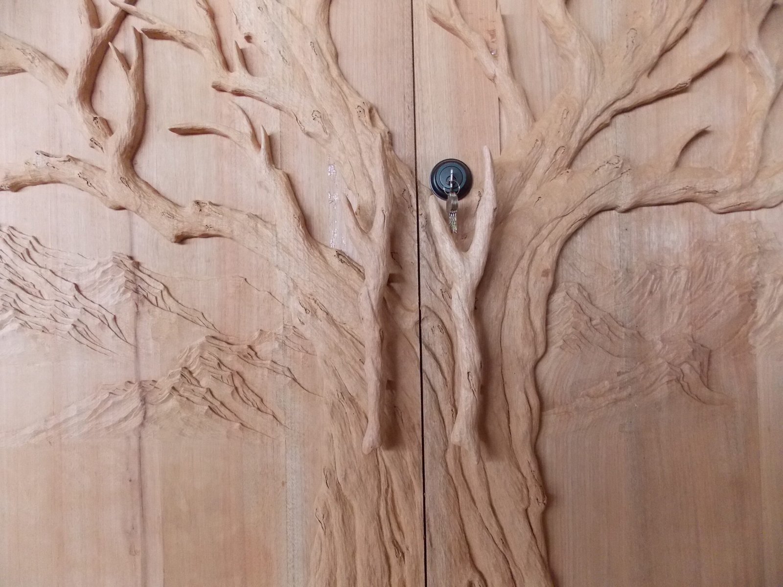 Centered view of hand-carved solid wood double entry door by Crown Custom Doors showing a prominent tree trunk split across two door panels, custom branch pulls, keyhole hardware, and mountain relief background.