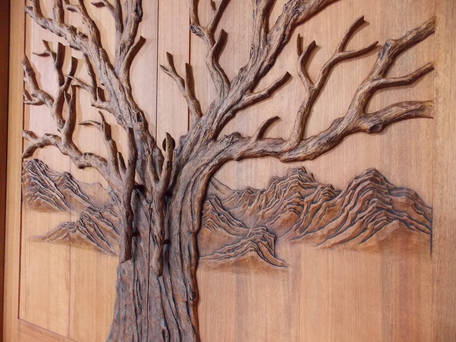 Angled close-up of a finished solid wood double entry door by Crown Custom Doors, showcasing deeply carved Tree of Life branch layers, custom branch pull handles, rich wood stain, and textured mountain peaks.