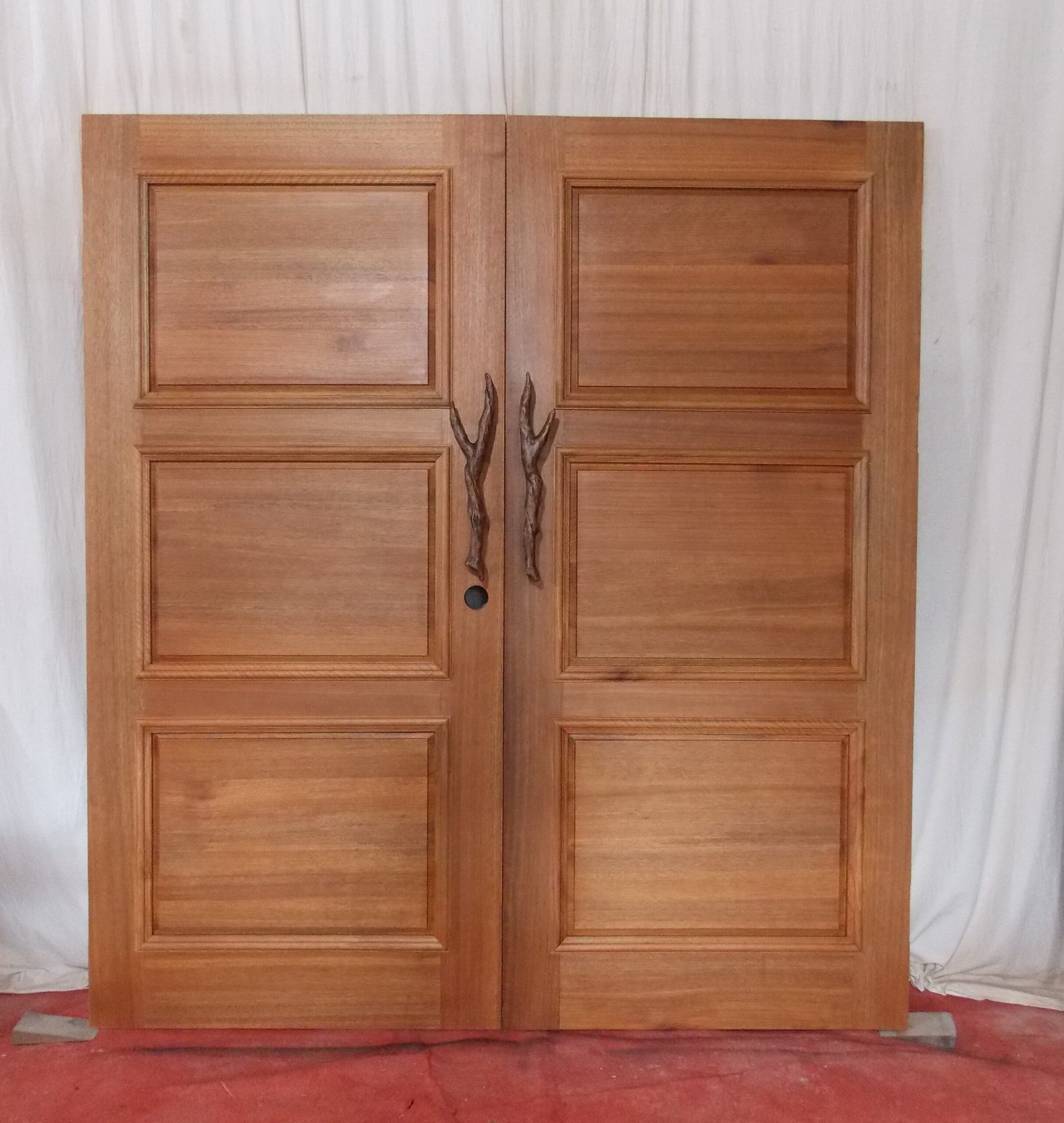 Studio photo showing the finished interior side of a stained solid wood double entry door by Crown Custom Doors, featuring classic three-raised-panel construction and custom hand-carved branch pull handles.