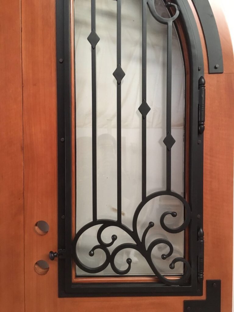 A close-up workshop view of a custom solid wood arched door by Crown Custom Doors, showcasing an operational matte black wrought iron window grille with elegant scrolls and diamond accents fitted with heavy-duty hinges and a spring latch over frosted privacy glass.