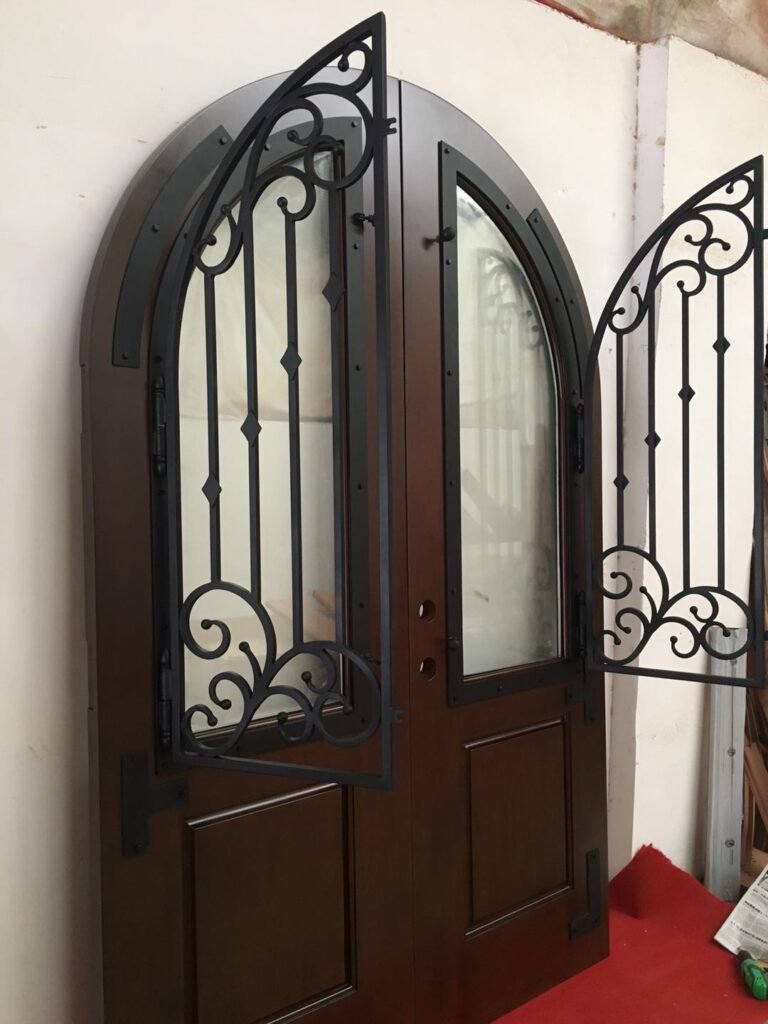 A custom arched solid wood double entry door system by Crown Custom Doors, showcasing both hand-forged black wrought iron window grilles swung completely open on their hinges away from the glass panels inside a workshop staging area.