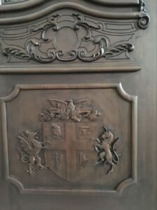 Front-facing close-up of a custom solid wood door panel showing a hand-carved heraldic family crest with a dragon and unicorn holding a shield, positioned beneath an elegant hand-carved baroque cartouche scrollwork frame.