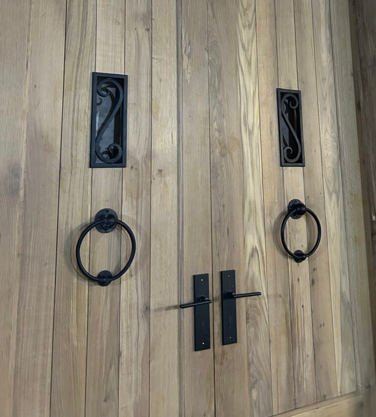 A close-up view of a custom double wood entry door showcasing a natural wood grain plank texture accented with two black wrought iron speakeasy grilles, matching heavy-duty iron ring pulls, and modern rustic black door handles.