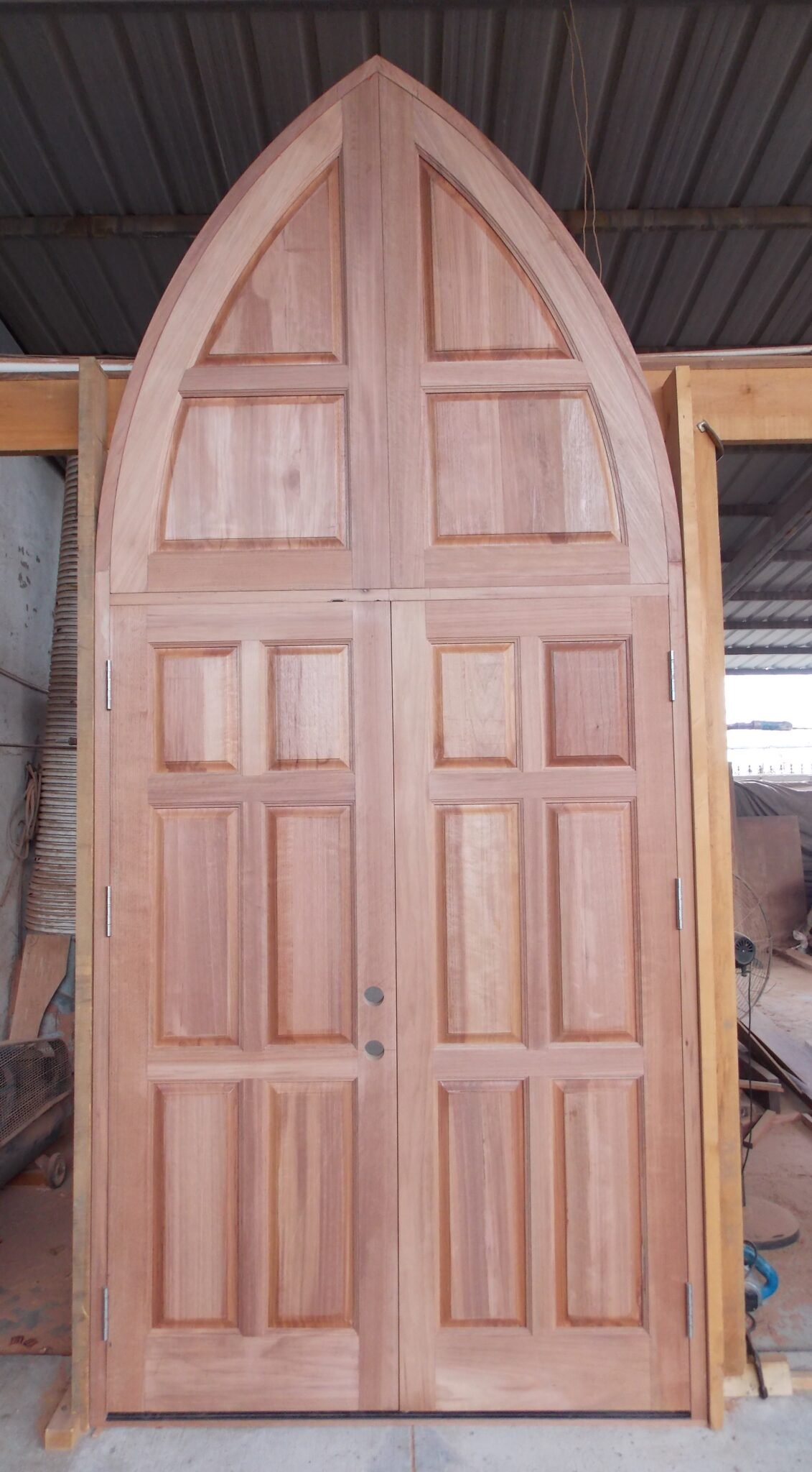 A raw, unfinished prehung Gothic pointed arch double entry door system custom-built from solid wood by Crown Custom Doors, shown fully assembled with hinges, pre-drilled boreholes, and a matching monumental pointed arch transom panel unit inside the workshop.