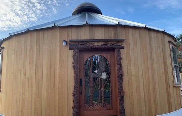 Full exterior view of a custom hand-carved solid mahogany bald eagle front door with tree branch casing installed on a round wooden yurt with a central roof dome.