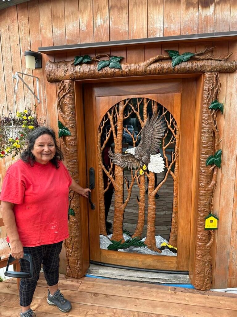 Happy client standing beside an installed custom hand-carved solid mahogany bald eagle front door with custom carved tree branch casing on a wooden yurt.