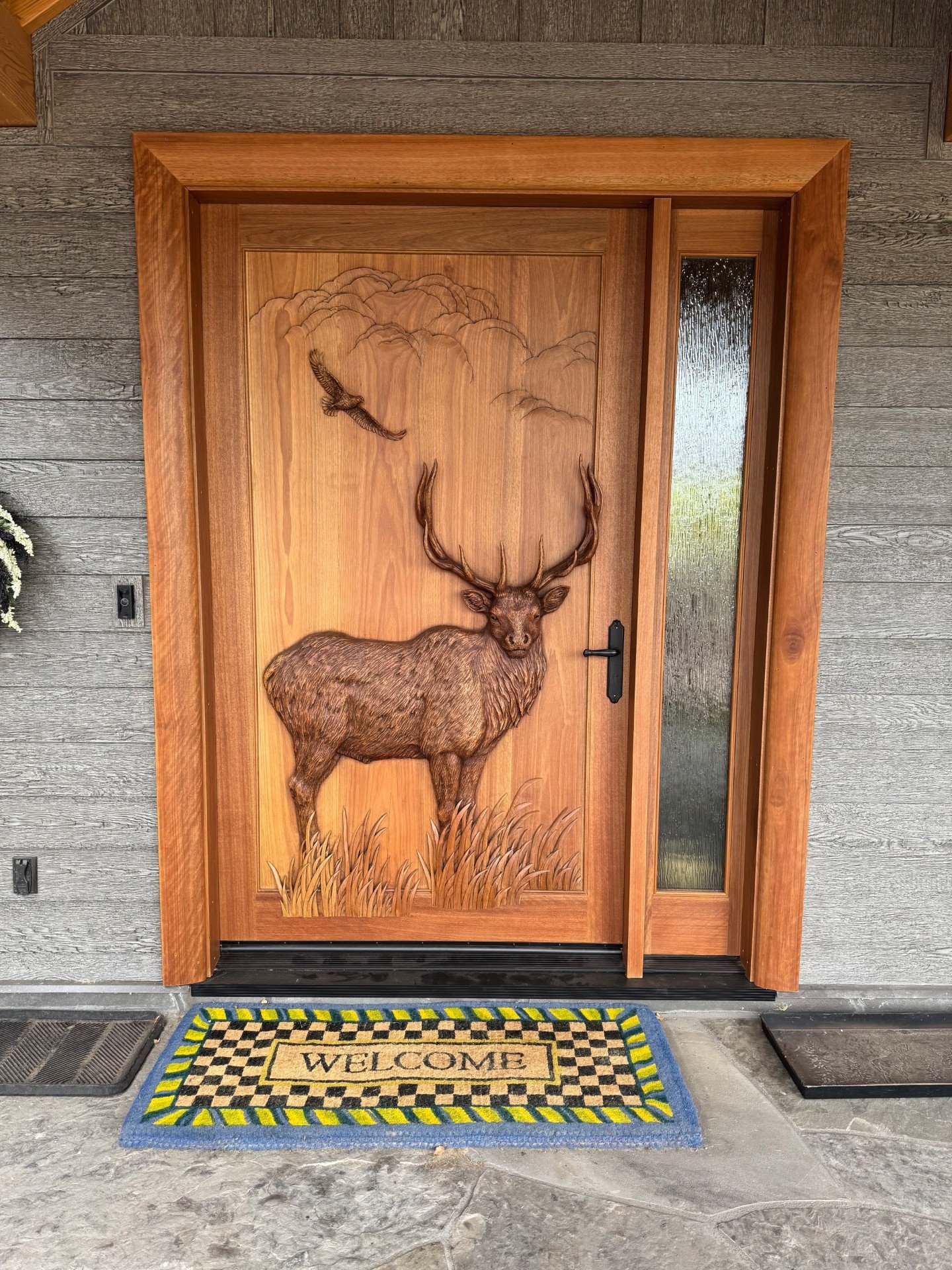 Installed custom solid wood single entry door with a hand-carved bull elk and soaring eagle relief paired with a textured privacy glass sidelight on a wood-sided home.