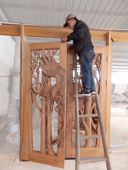 Craftsman standing on a ladder in a woodworking workshop, testing alignment on pre-hung hand-carved solid wood eagle double doors.
