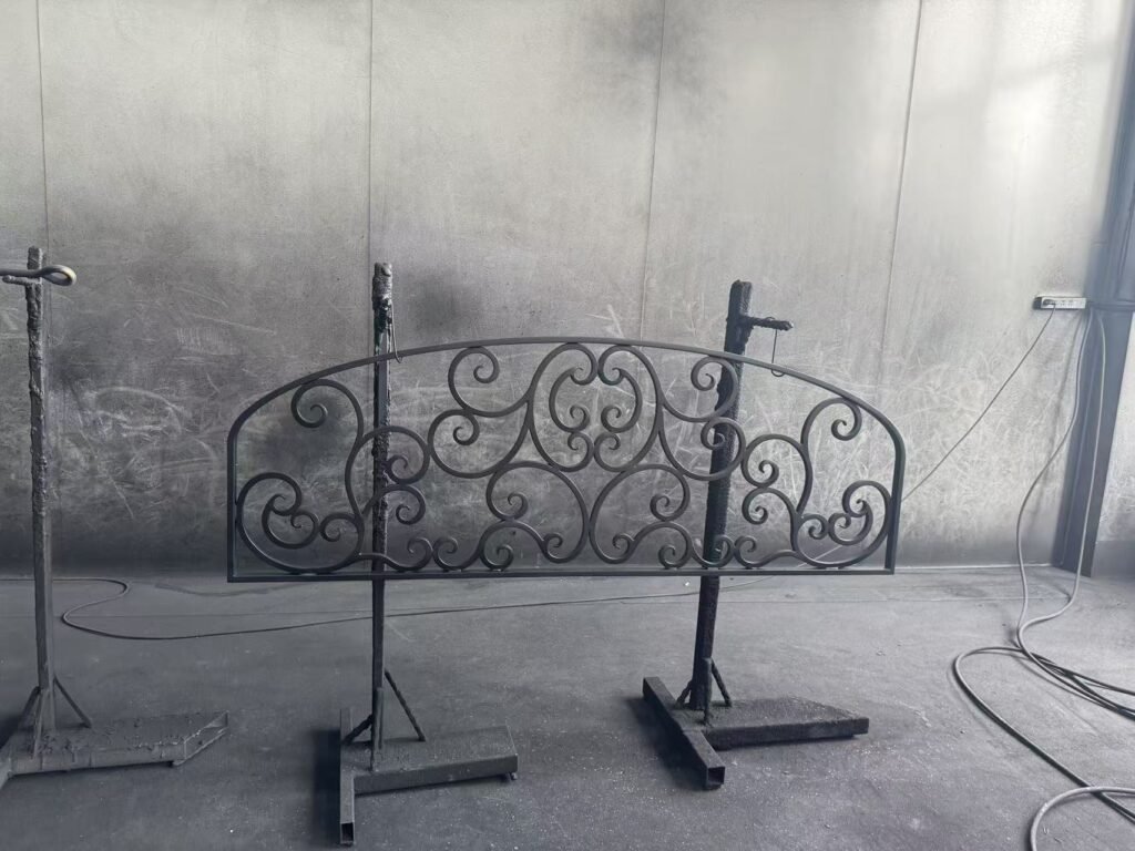 A hand-forged black wrought iron arched transom door grille featuring elegant symmetrical scrollwork, shown resting on metal stands inside a workshop finishing booth during production.