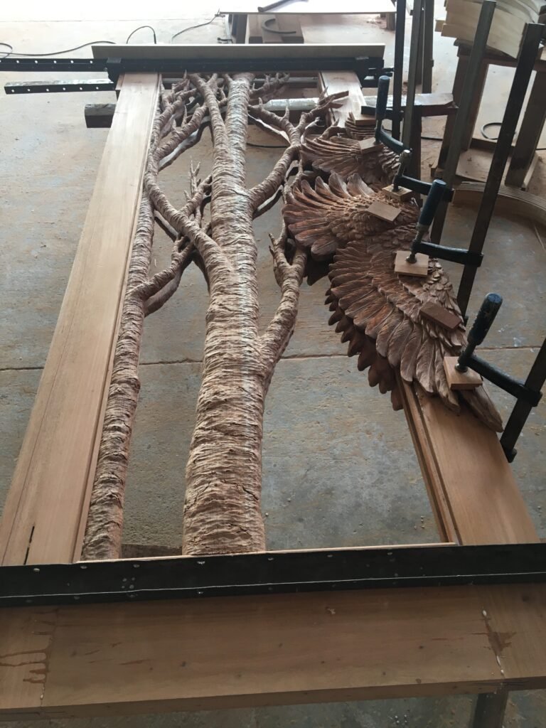 Full solid wood door frame clamped on a workshop workbench during assembly, featuring mounted hand-carved tree trunks, branches, and eagle wings.