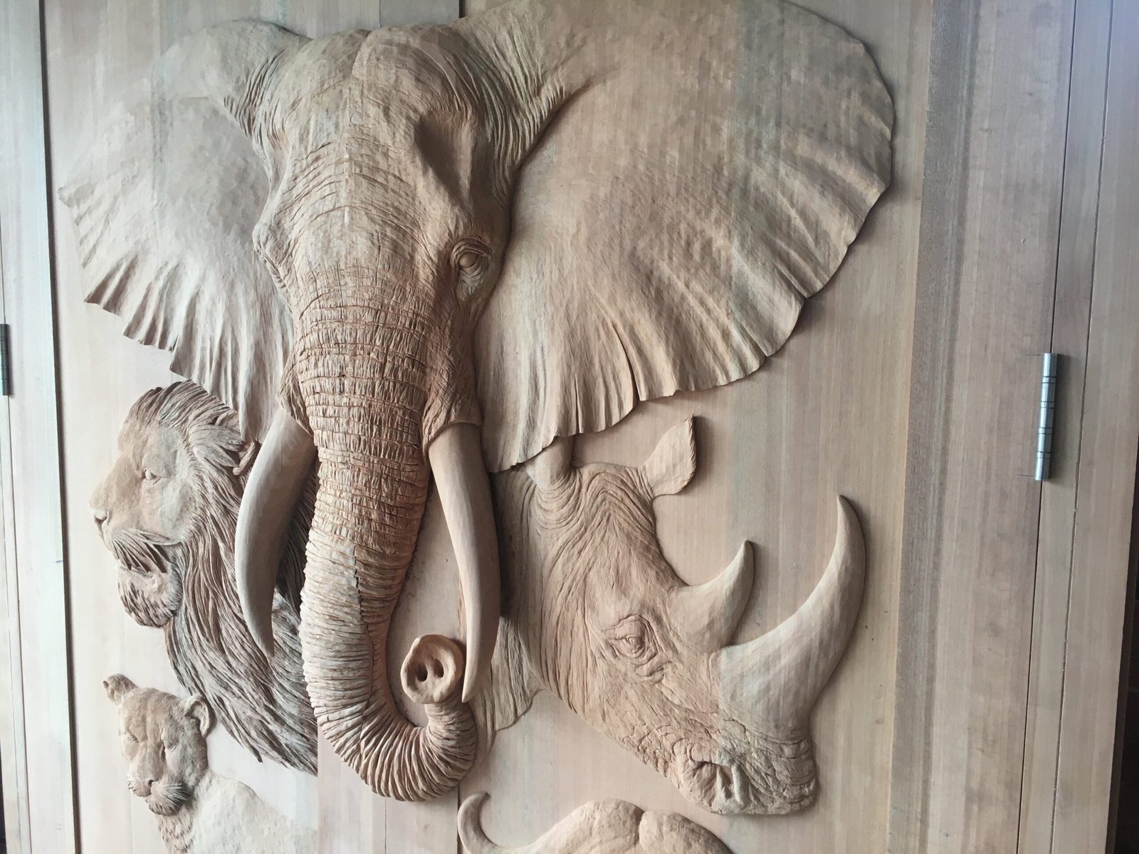 A close-up, three-quarter workshop view of a raw solid wood double door highlighting the deep high-relief hand carvings of an African elephant head, a roaring lion, a rhinoceros head with prominent horns, and a leopard.