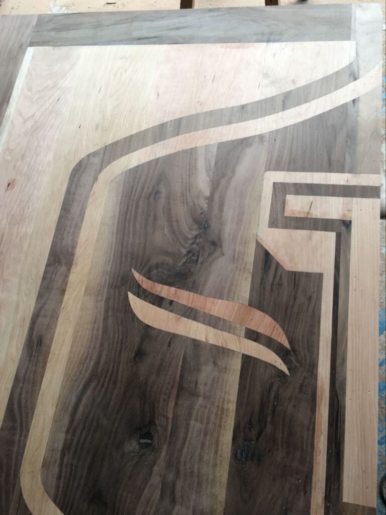 A close-up view of an unfinished solid wood door panel showing the intricate grain patterns and flush joints of a dark black walnut shield and light cherry wood inlay.