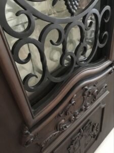 Angled close-up of a custom solid wood entry door showing a hand-forged black wrought iron scrollwork grille over a glass insert, set above deep, hand-carved baroque wood moldings and a coat of arms panel.