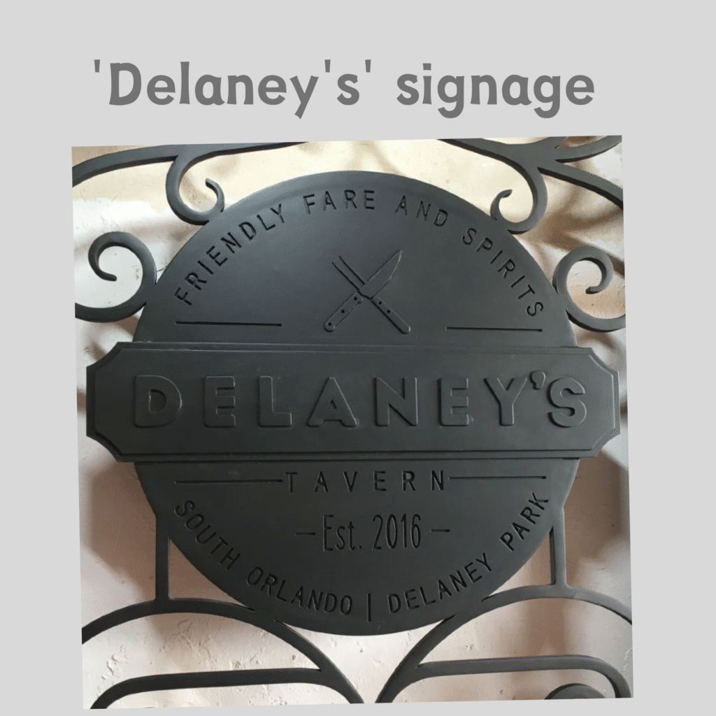 An up-close view of a custom matte black wrought iron restaurant signage medallion integrated into a door grille by Crown Custom Doors, showcasing embossed lettering for "Delaney's Tavern — Est. 2016 — Friendly Fare and Spirits — South Orlando | Delaney Park" paired with a crossed knife and fork icon surrounded by hand-forged scrollwork.