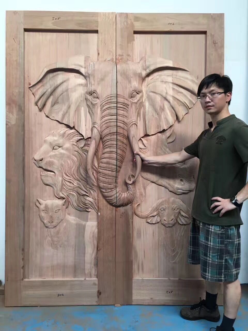 The master designer artisan stands next to a massive, raw solid wood double door in a workshop, placing his hand on the high-relief carving of an African elephant tusk. The door also features detailed hand-carved reliefs of a lion, rhinoceros, leopard, and cape buffalo.