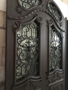 Angled close-up view of a custom hand-carved solid wood double door, showcasing the deep 3D relief of the moldings, a hand-carved leaf motif, and heavy-duty hand-forged black wrought iron scrollwork over glass.