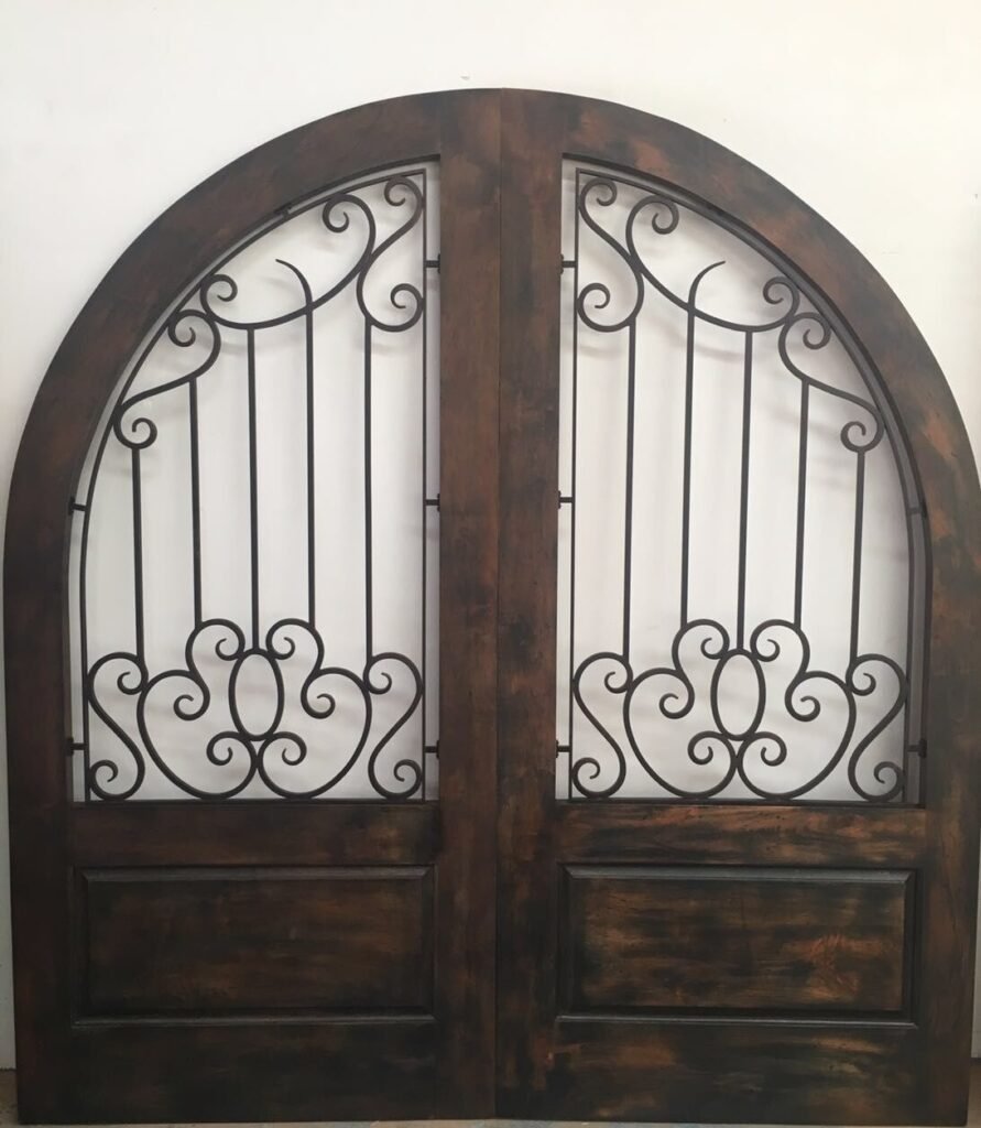 A completed full-arched solid wood double door set by Crown Custom Doors, finished in a rich dark brown custom stain with intricate hand-forged black wrought iron scrollwork grilles over the glass panels.