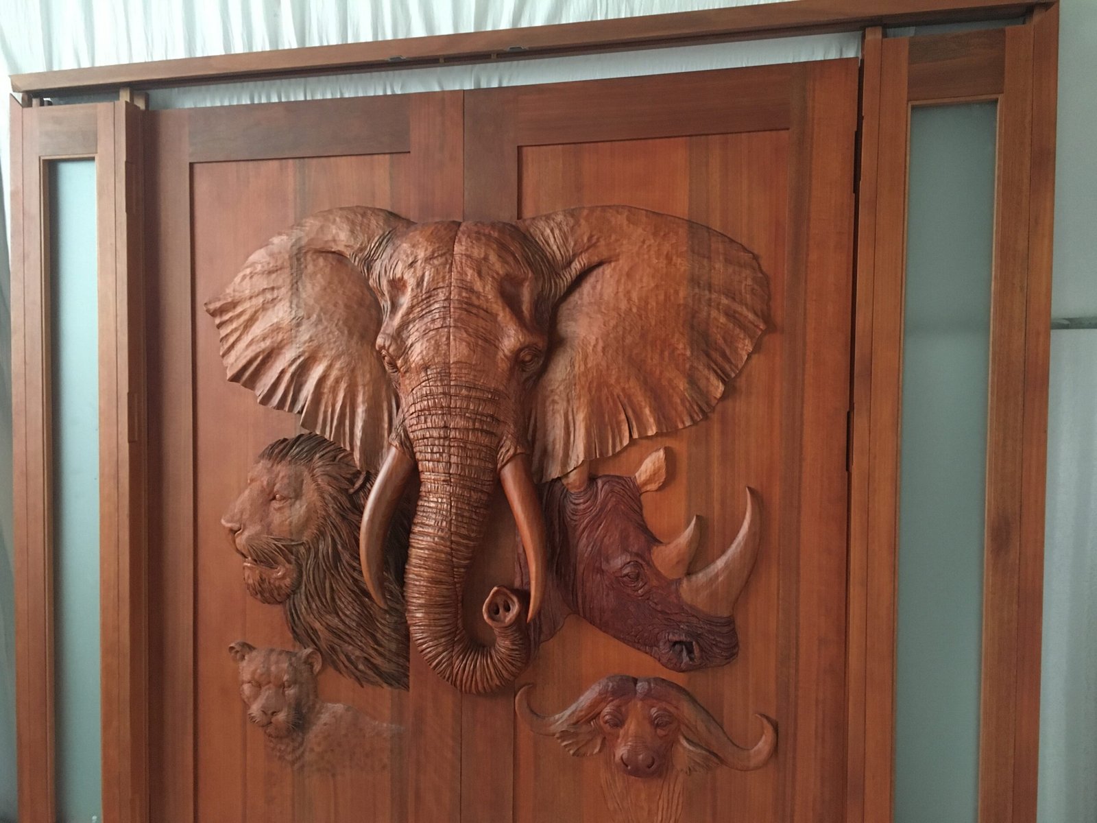 A front view of a finished, stained solid wood double entry door featuring 3D high-relief carvings of an African elephant, lion, rhinoceros, leopard, and cape buffalo, assembled with matching wooden sidelights containing frosted glass.