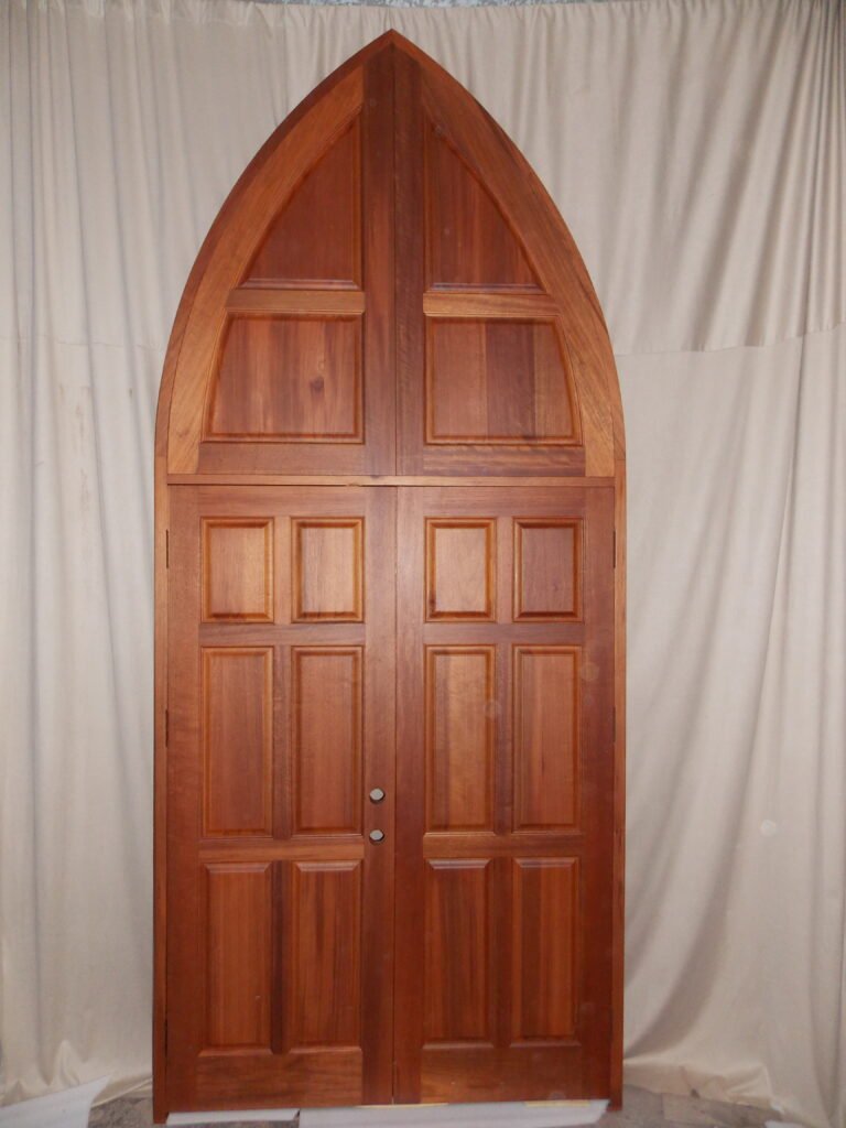 A finished custom Gothic pointed arch double entry door system crafted from solid wood by Crown Custom Doors, featuring a rich satin amber stain, a matching grand multi-panel pointed arch transom unit, integrated side hinges, and pre-drilled hardware bores staged against a neutral backdrop.