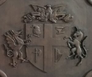 Close-up of a custom hand-carved heraldic coat of arms on a solid wood door, featuring a detailed central shield with a cross, a dragon supporter on the left, a unicorn supporter on the right, and a carved eagle crest on top.