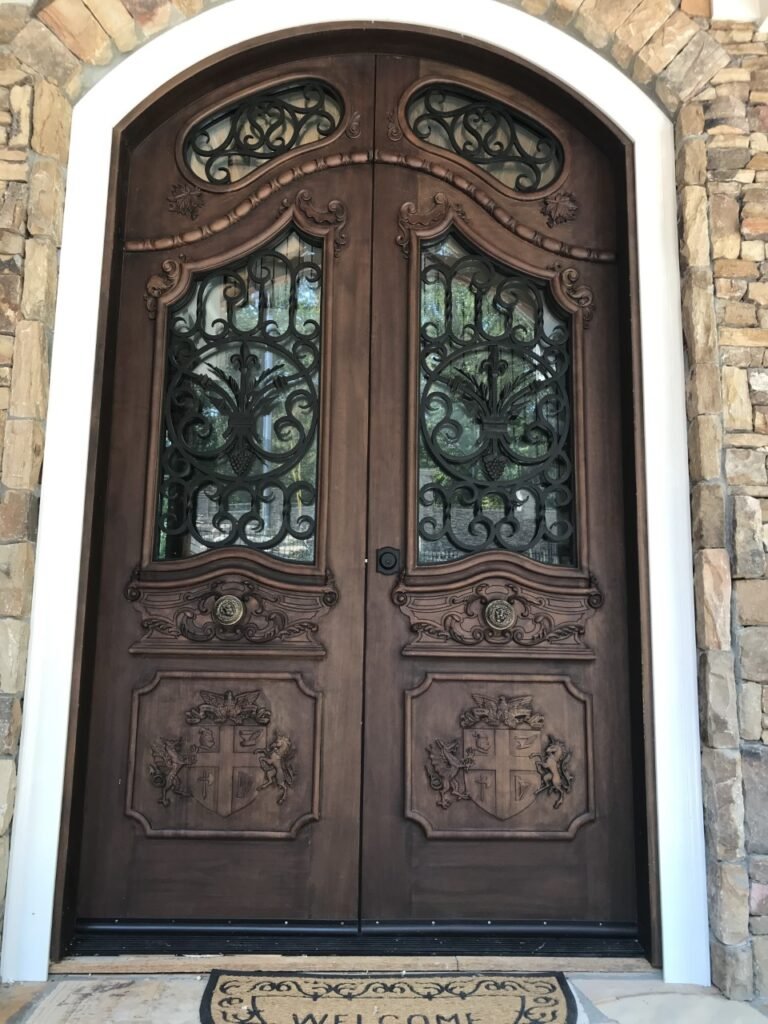 Ornate, custom-carved solid wood double entry door featuring highly detailed hand-carved family crests on the lower panels, classical high-relief moldings, and decorative black wrought iron scrollwork over glass panels.