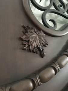 Close-up of a custom-carved solid wood door panel showing high-relief, hand-carved leaves, wheat stalks, and a pinecone detail next to a curved arched trim and a black wrought iron window insert.