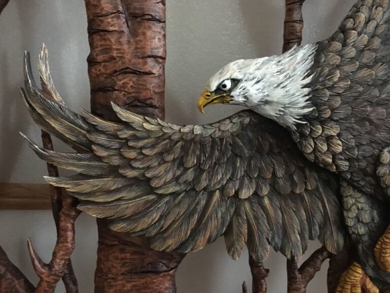 Close-up of the painted outstretched wing and white head of a hand-carved mahogany bald eagle with a carved birch tree trunk in the background.
