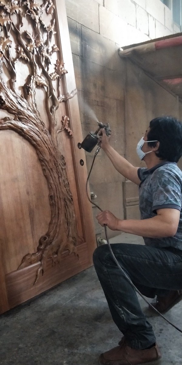 Woodworking craftsman in a workshop applying a protective topcoat spray finish to a hand-carved solid wood Eternal Oak tree door slab.