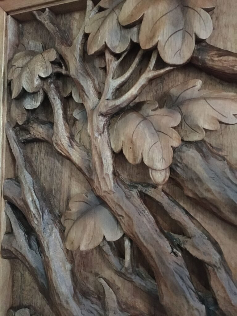 Close-up view of a hand-carved solid wood panel showcasing detailed oak tree branches, carved oak leaves, and acorns in high relief.