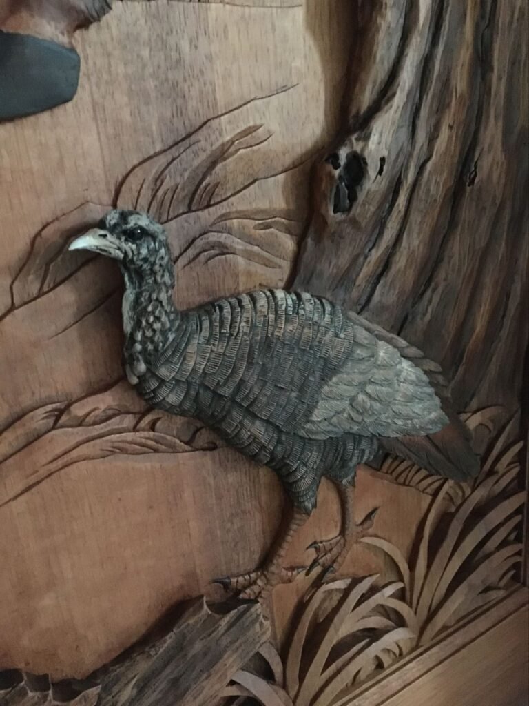 Close-up view of a hand-carved and hand-painted wild turkey standing near grass and tree bark on a solid wood door panel.