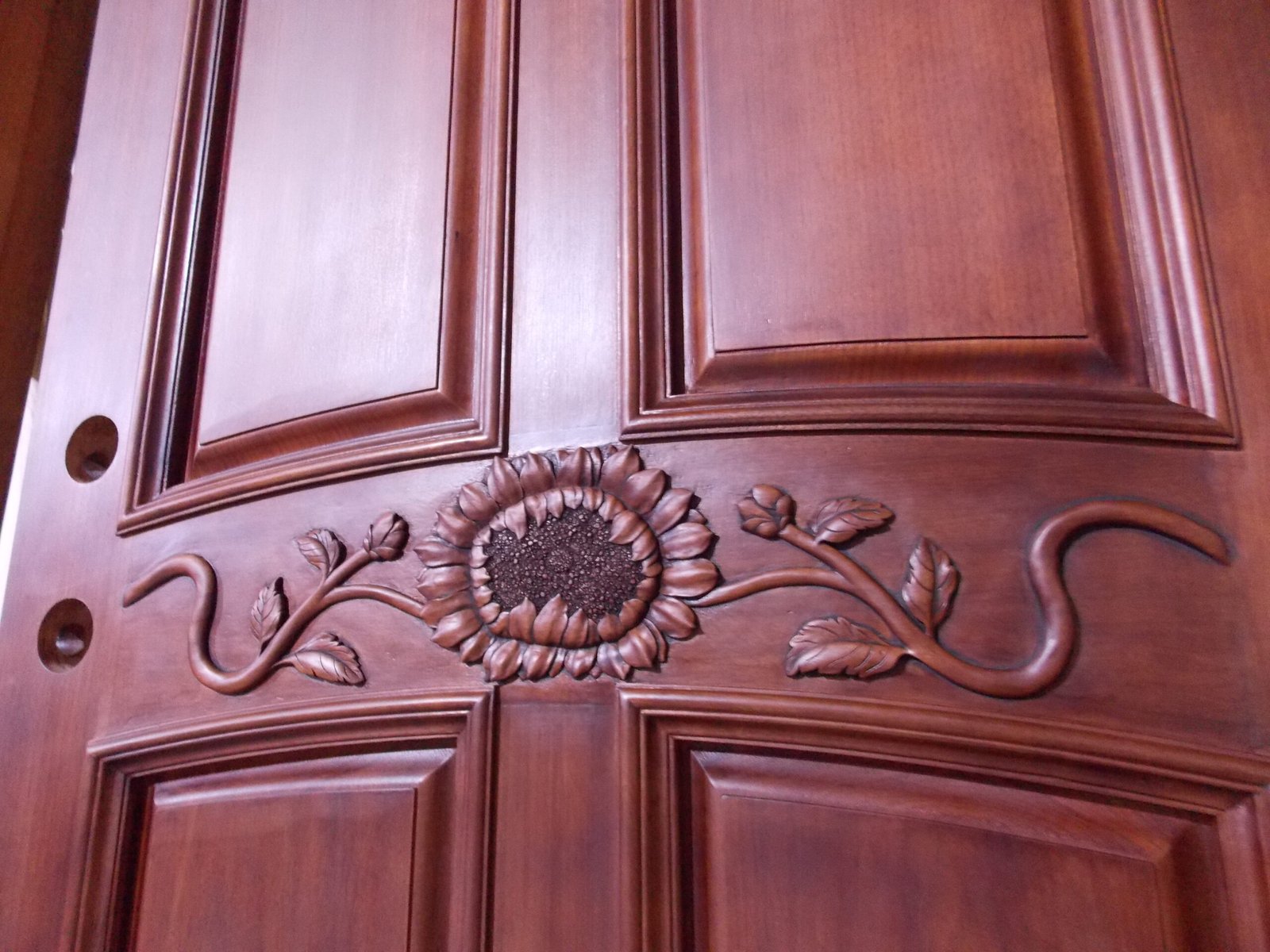 Macro close-up of a central hand-carved sunflower and vine design on a premium solid mahogany wood front door, showing the intricate carving detail, rich finish, and raised panel construction.
