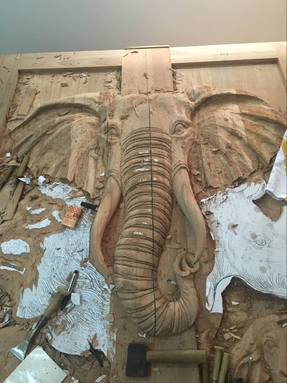 A top-down view of a custom solid wood double door in a workshop during the hand-carving process, showing a deeply sculpted high-relief African elephant head surrounded by wood shavings, traditional hand chisels, a mallet, and paper layout templates