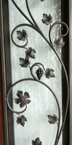 Close-up of a custom handcrafted front door featuring an ornate wrought iron grapevine design with detailed leaves and a grape cluster over textured privacy glass.