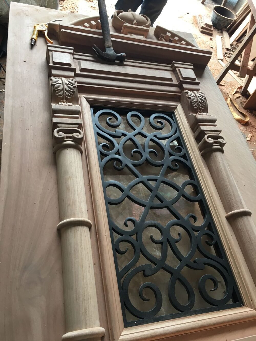 A behind-the-scenes look inside the Crown Custom Doors workshop, showing a craftsman assembling a solid wood Gothic Revival front door panel on a workbench, highlighting the integration of hand-carved pillars, ornate pediment trim molding, and a custom black wrought iron scrollwork grille.